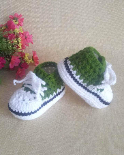 Hand-knitted baby bootees in green and white with lace detail, displayed on a beige surface next to pink flowers.